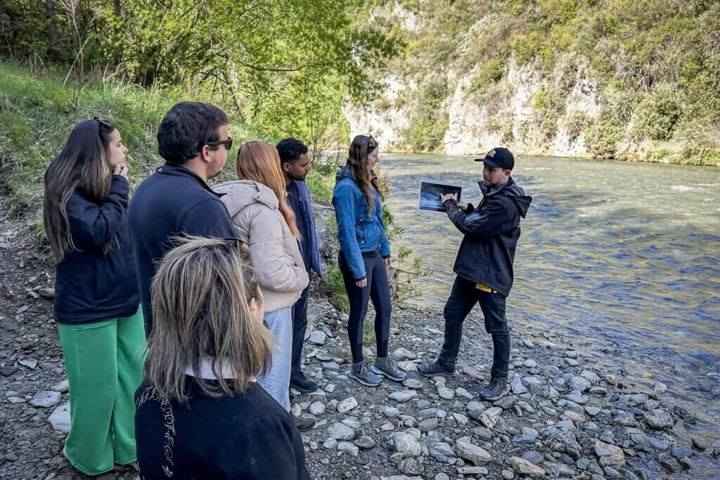 Tour guide at river with tourists explaining a film scene with a printout from the movie 