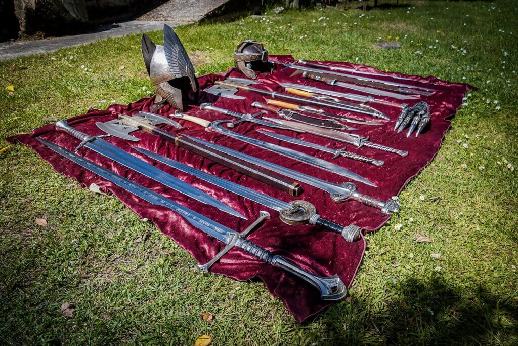 A variety of different prop swords, helmets and axes layed out on a red velvet sheet